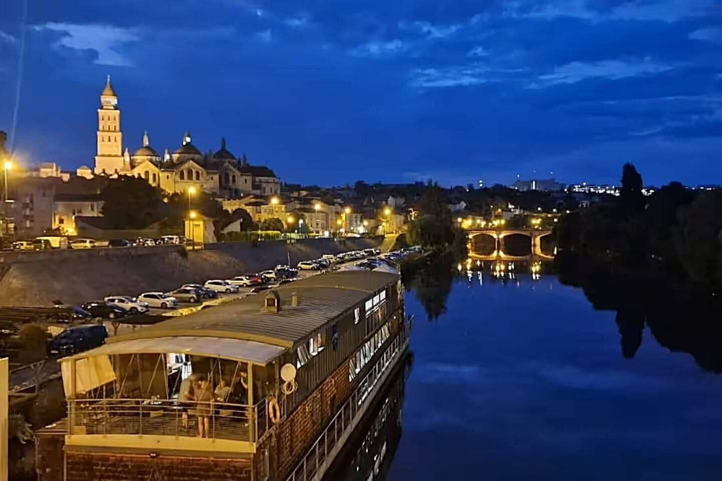 Une rivière avec un bateau amarré sur la rive gauche, un pont au milieu et un bâtiment avec une tour sur la rive droite.