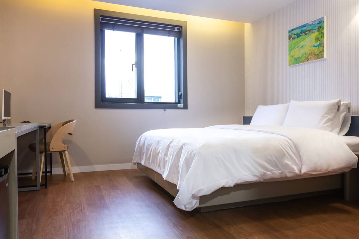 Ein Schlafzimmer mit einem Bett, einem Schreibtisch mit Computer, einem Sessel und einem Fenster.