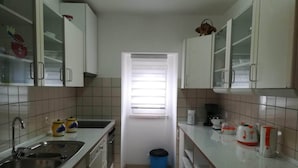 A compact kitchen with white cabinets, a stainless steel sink, a blue trash bin, and a window with blinds. - Casa da Avó near the beaches of the western zone (Óbidos)