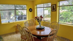 A dining area with a round wooden table surrounded by six white chairs, yellow walls, and large windows offering a view of the outdoors. - 2 bedroom cottage just a short walk to Swan River (Dennis Port)