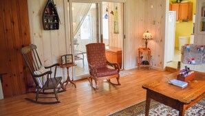 A cozy living room with wooden flooring, a rocking chair, a patterned armchair, a small table with a lamp, and a colorful rug. - 2 bedroom cottage just a short walk to Swan River (Dennis Port)