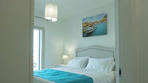 A bedroom with a bed, a nightstand, a lamp, a painting of a coastal scene, and a window.