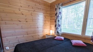 Una habitación con paredes de madera, una cama con ropa oscura y dos almohadas rosas, una mesita de noche con una lámpara y una ventana con cortinas florales.