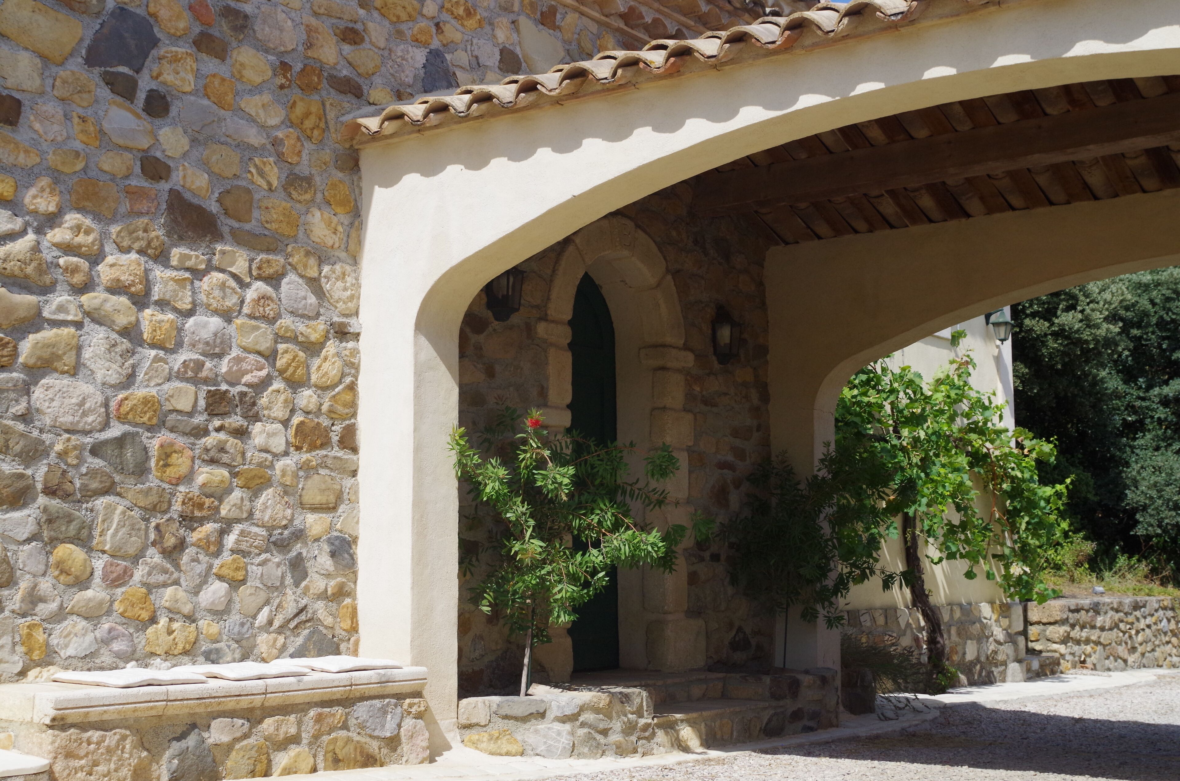 A stone building with a tiled roof and an arched entrance. There are steps leading up to the entrance, and a small tree with green leaves is visible.