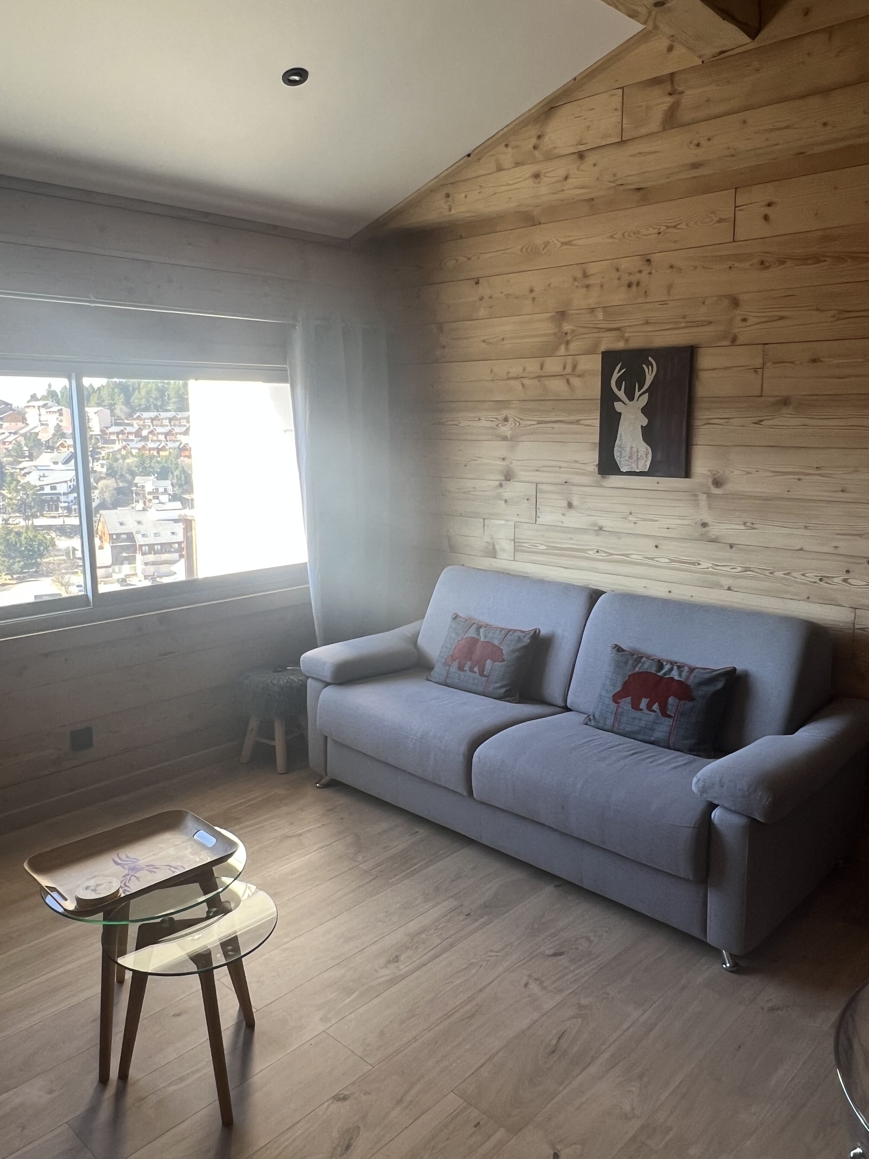 A living room with a grey sofa, two bear-themed pillows, a glass coffee table, and a wooden wall with a deer painting.