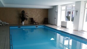 Ein Hallenbad mit klarem, blauem Wasser, umgeben von gefliestem Boden und weißen Wänden. In der Nähe der Wand befinden sich zwei Liegestühle und ein Topfpflanze.