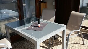 Ein Tisch mit Glasplatte, zwei Stühle, eine Flasche und zwei Weingläser auf einer Terrasse mit Blick auf ein Gebäude und eine Tür.