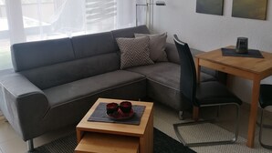 Ein modernes Wohnzimmer mit einer grauen Eckcouch, einem Holztisch, einem schwarzen Bürostuhl und einem kleinen Holzbüro mit einer Lampe. An der Wand hängen zwei Landschaftsbilder und durch große Fenster ist die Außenwelt zu sehen.