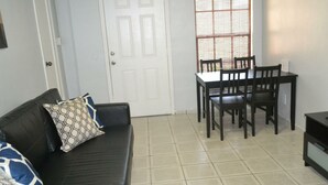 Uma sala de estar com um sofá preto, almofadas estampadas, uma mesa de jantar com cadeiras e uma porta branca.