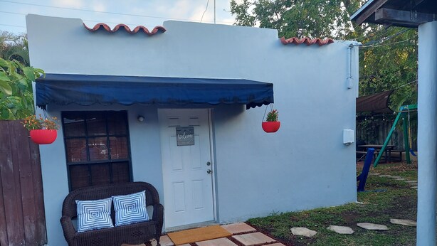 Uma pequena área externa privativa com toldo azul, uma cadeira de vime com almofada listrada e uma placa de boas-vindas na porta.