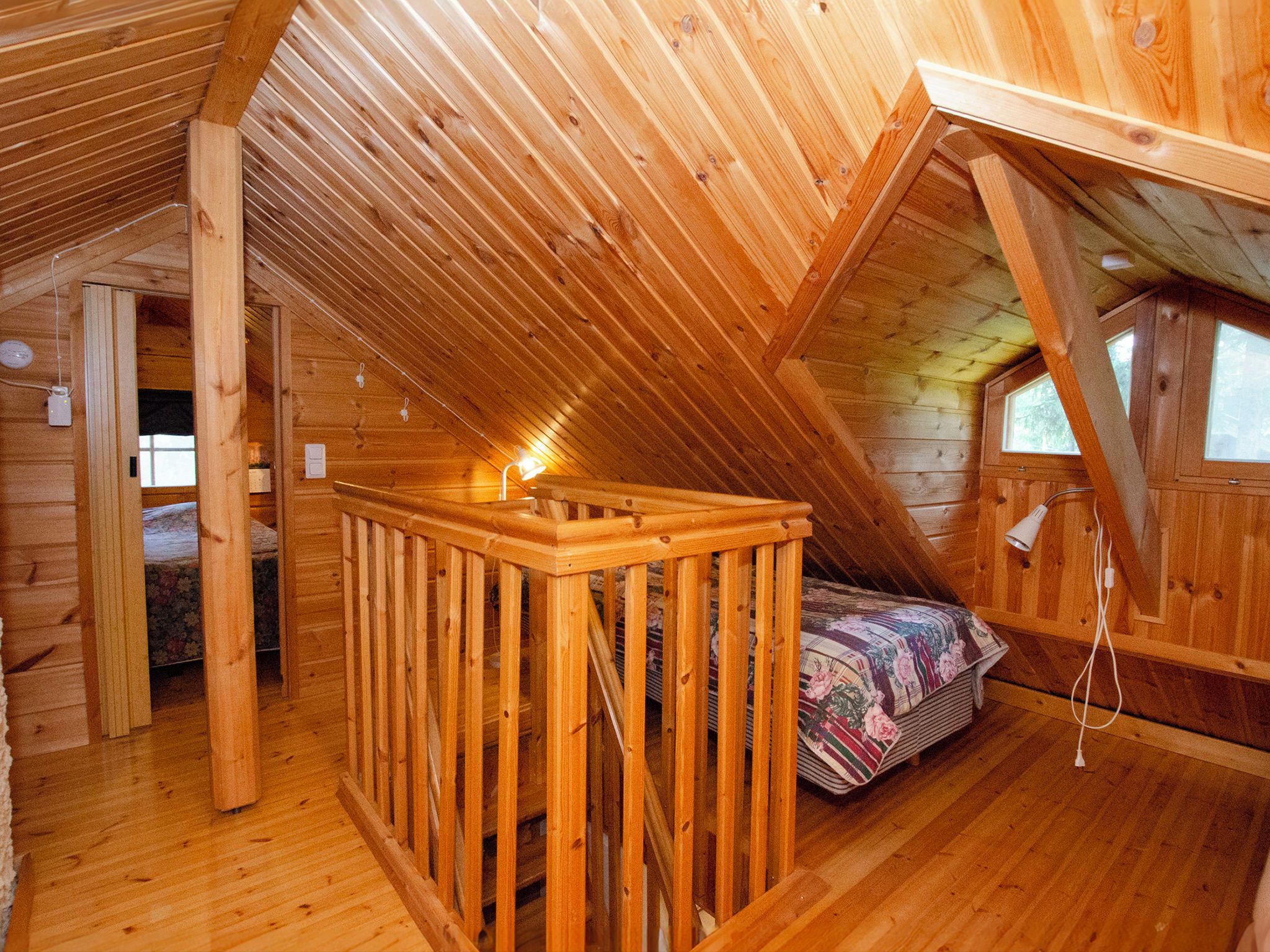 A wooden attic bedroom with a sloped ceiling, a wooden railing, a bed with a floral bedspread, and a window.