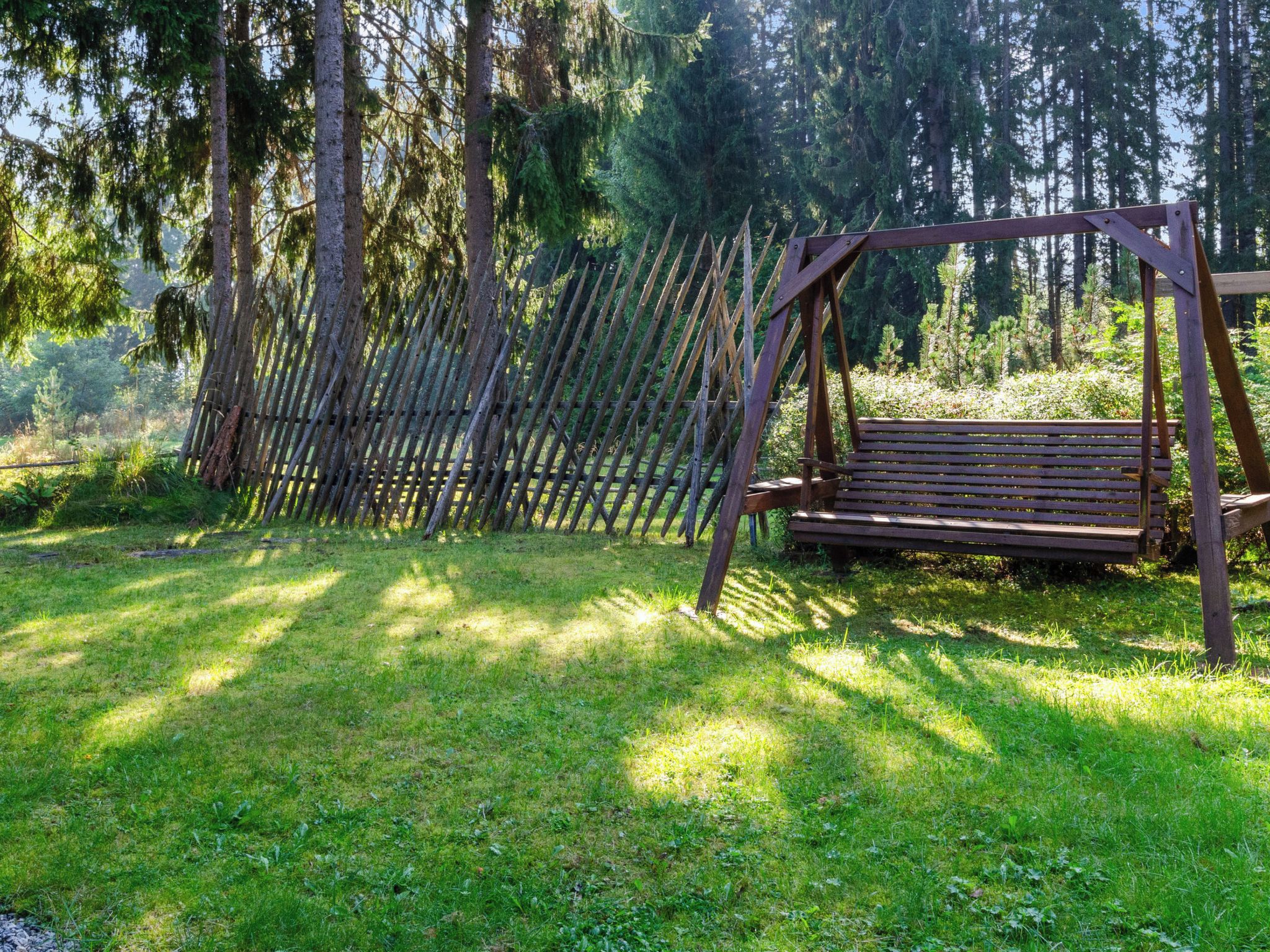 Un ensemble de balançoires en bois installé dans une zone herbeuse, avec une clôture en bois et des arbres en arrière-plan.