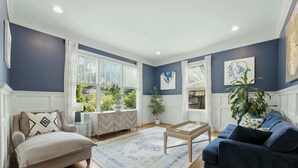 Ein Wohnzimmer mit Sofa, Sessel, Couchtisch und einem großen Fenster mit Vorhängen.