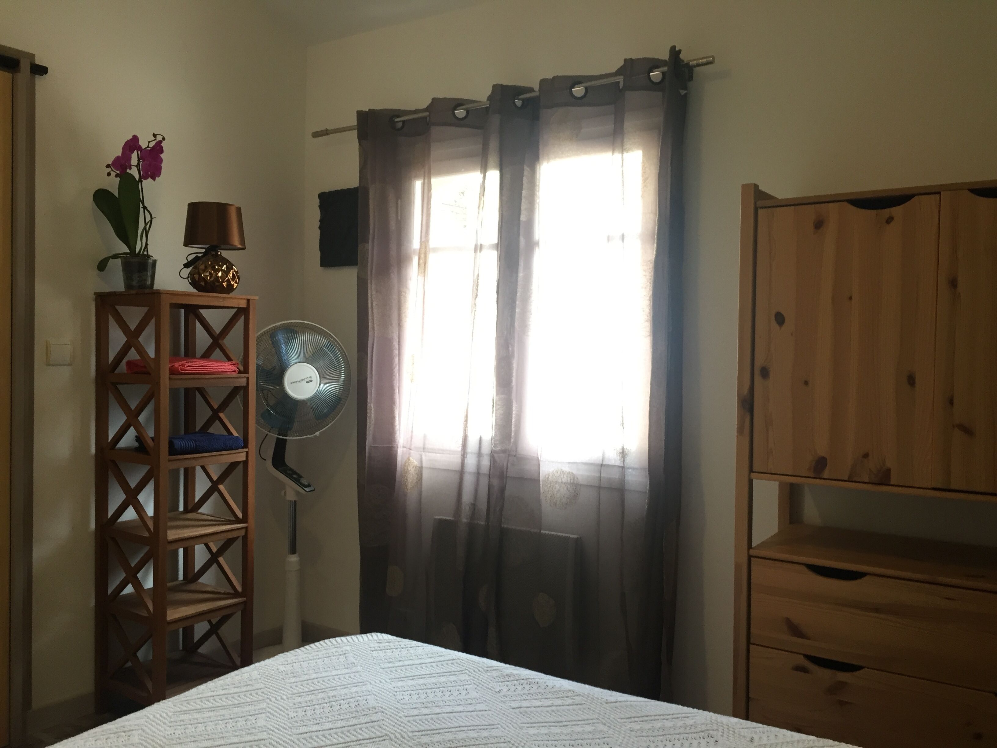 Une chambre à coucher avec un lit, une armoire en bois, un ventilateur sur pied, une étagère avec des serviettes et un pot contenant une orchidée.