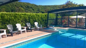 Une piscine entourée d’une structure en verre, bordée de chaises Adirondack blanches, et offrant une vue sur une colline verdoyante.