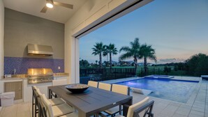 An outdoor kitchen with a grill, dining table, and chairs. - Modern Luxury Villa | 5BR, 5 Bath Luxe Home w/ Private Pool, Arcade, and Theater Near Disney (Kissimmee)