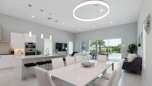 A modern kitchen with white cabinetry, a central island with bar stools, and a dining area with a large table and chairs. - Modern Luxury Villa | 5BR, 5 Bath Luxe Home w/ Private Pool, Arcade, and Theater Near Disney (Kissimmee)