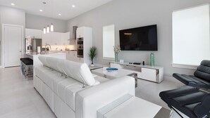 A modern living room with a white sofa, a black leather chair, a TV mounted on the wall, and a kitchen area in the background. - Modern Luxury Villa | 5BR, 5 Bath Luxe Home w/ Private Pool, Arcade, and Theater Near Disney (Kissimmee)