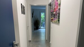 Ein Flur mit einer blauen Tür, weißen Wänden und einem gefliesten Boden, der zu einem Zimmer mit einem großen Fenster und einer Pflanze führt.