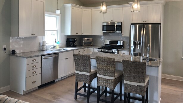 A modern kitchen with white cabinets, stainless steel appliances, and a central island with wicker chairs. - Walkable Location Cape May Home w Pool, 1st-Fl BR, Near Dining, Market & Beach (West Cape May)