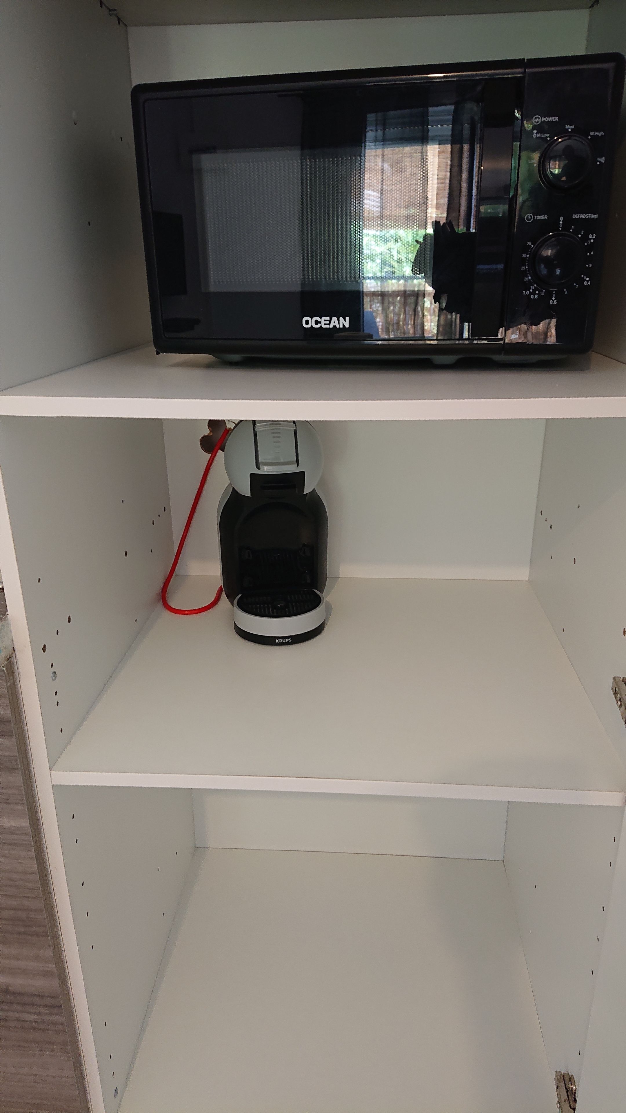 A black microwave on a white shelf, a Krups coffee maker below it, and a white wall.