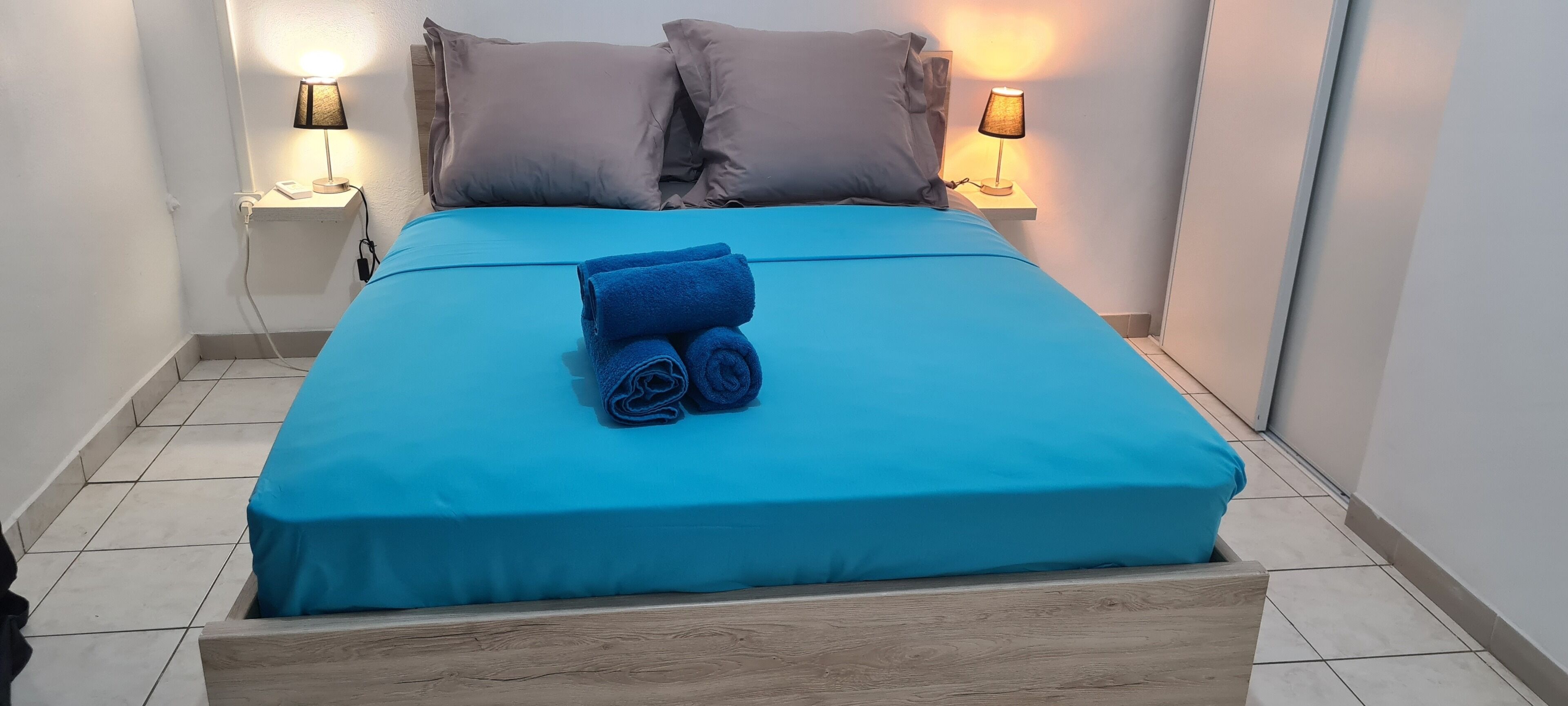 A neatly made bed with a blue bedspread and two rolled towels, two bedside lamps, and a wooden headboard.