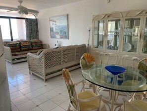 A living room with a glass dining table, wicker furniture, and a large window.