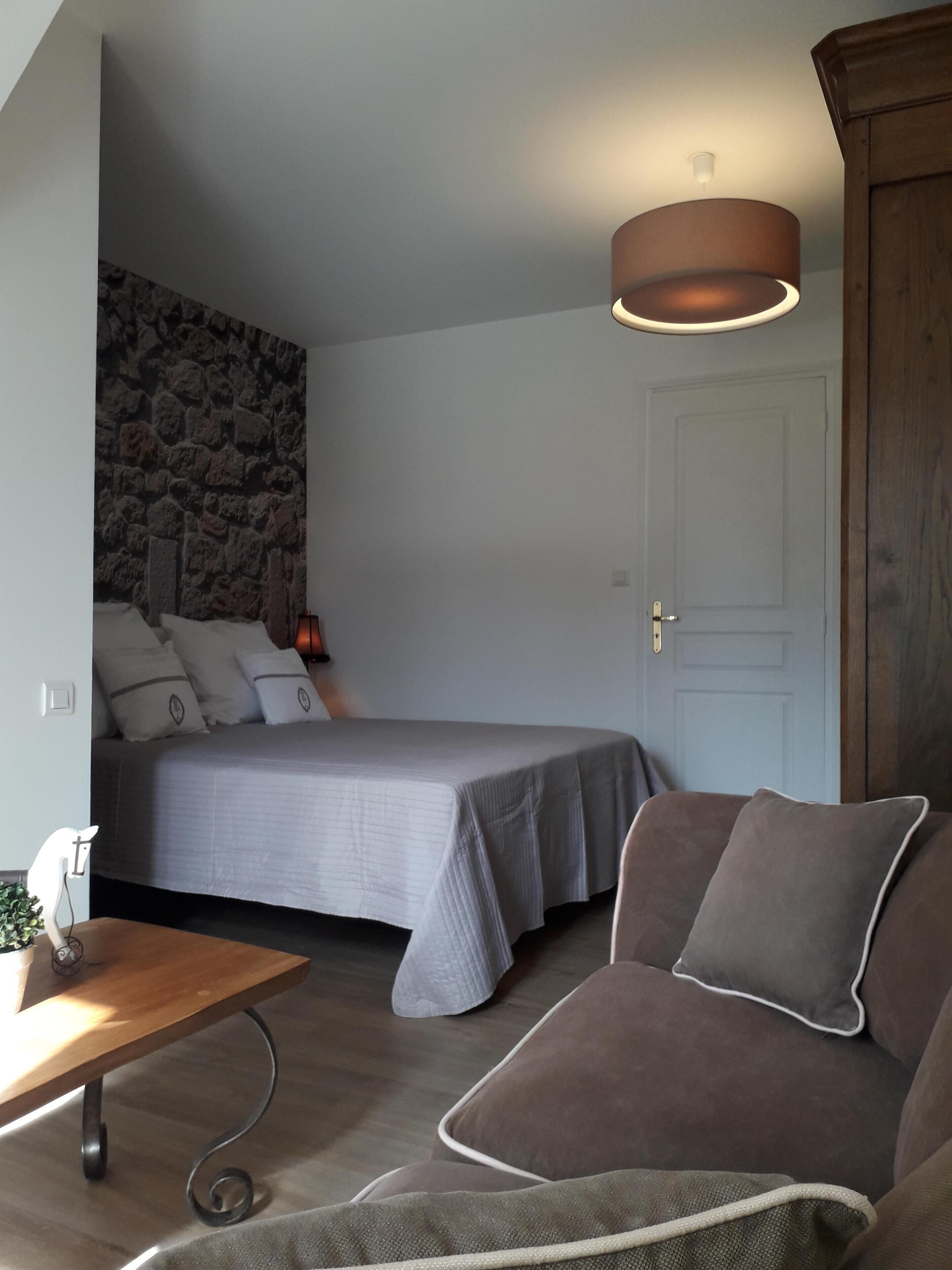 A bedroom with a bed, a wooden nightstand, a desk, a sofa, a stone wall, and a door.