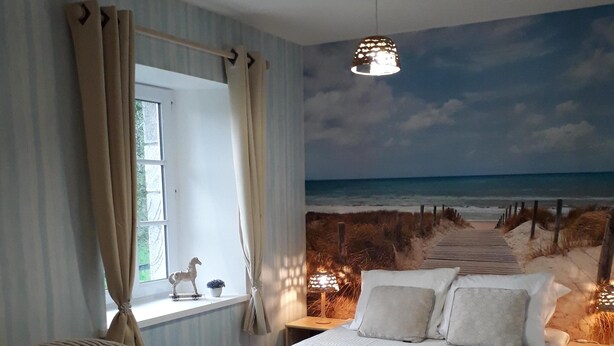 Ein Schlafzimmer mit einem Bett, einem Sessel, einem kleinen Tisch und einem Wandgemälde mit Strandmotiv.