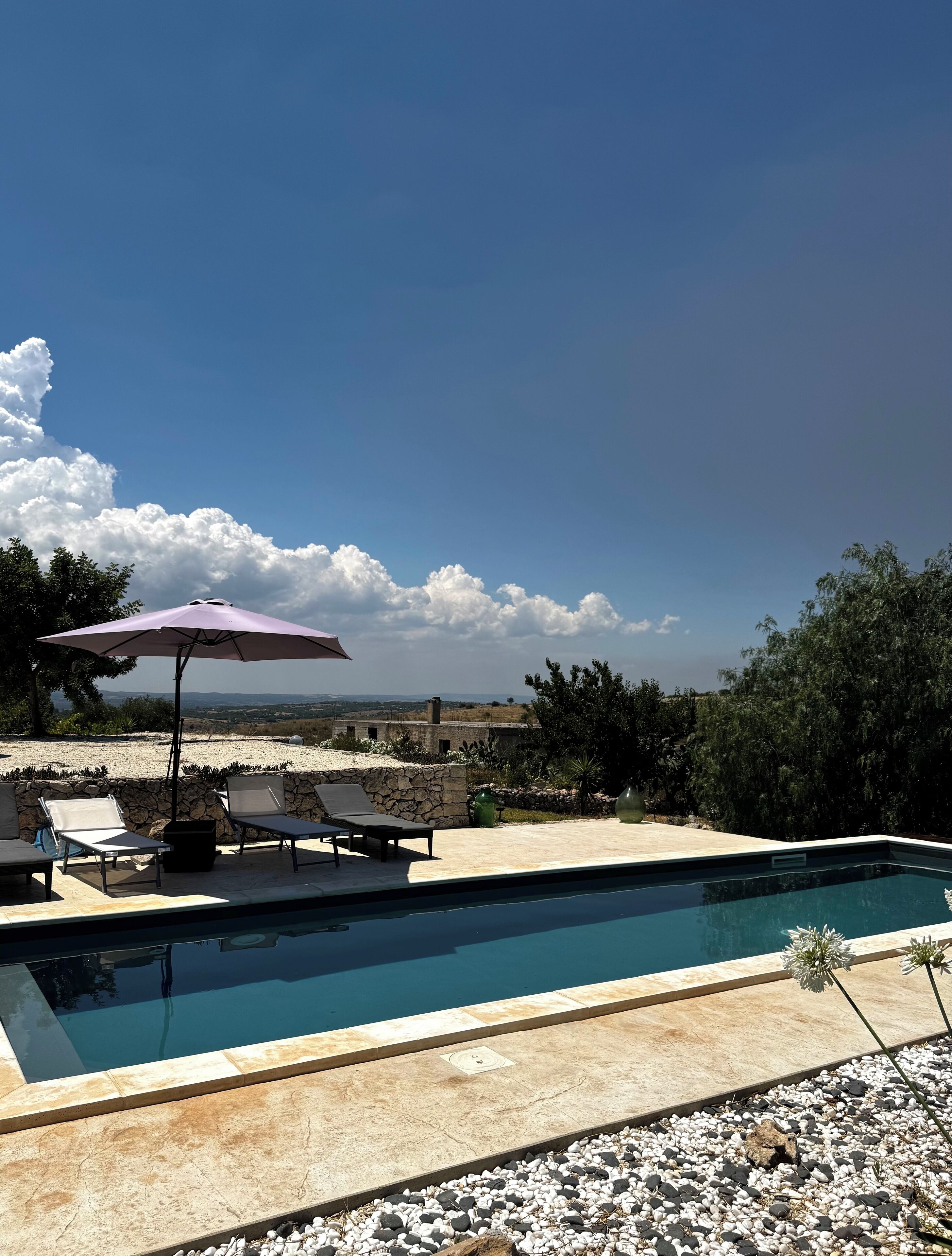 Une piscine entourée d’un mur de pierre et de verdure, agrémentée de chaises longues et d’un parasol, d’où l’on peut admirer les collines lointaines.