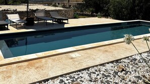 Une piscine entourée d’un mur de pierre et de verdure, agrémentée de chaises longues et d’un parasol, d’où l’on peut admirer les collines lointaines.