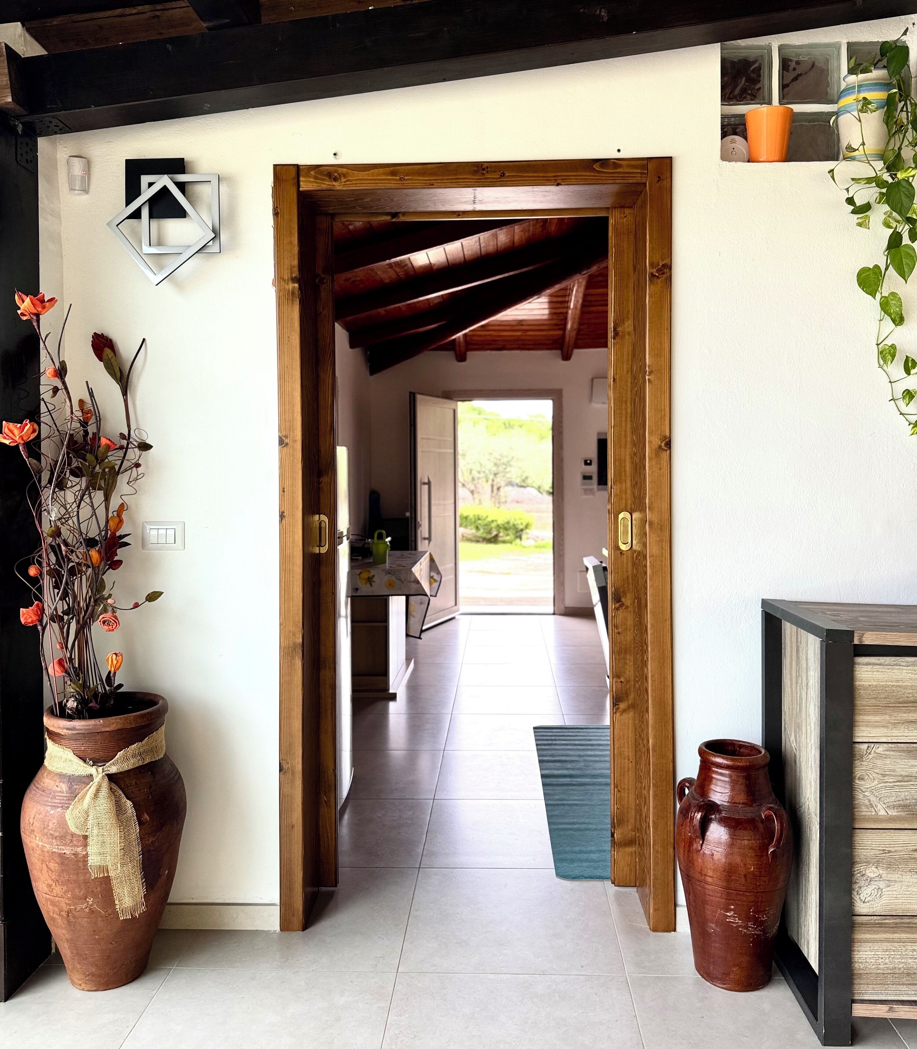 Un couloir avec une porte en bois donnant sur une pièce, une plante en pot et un vase posé sur le sol.