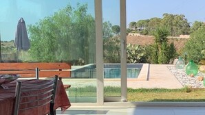 Un espace extérieur aménagé en terrasse, avec une table et des chaises, une porte vitrée donnant accès à une piscine, et une vue sur un jardin paysager avec des arbres et une colline.