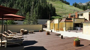 Una terraza de madera con sillones de descanso y sombrillas, un edificio de fachada amarilla y un paisaje montañoso cubierto de bosque al fondo.