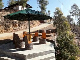 Un área con bancos de piedra, una sombrilla verde, rodeada de árboles y un edificio al fondo.