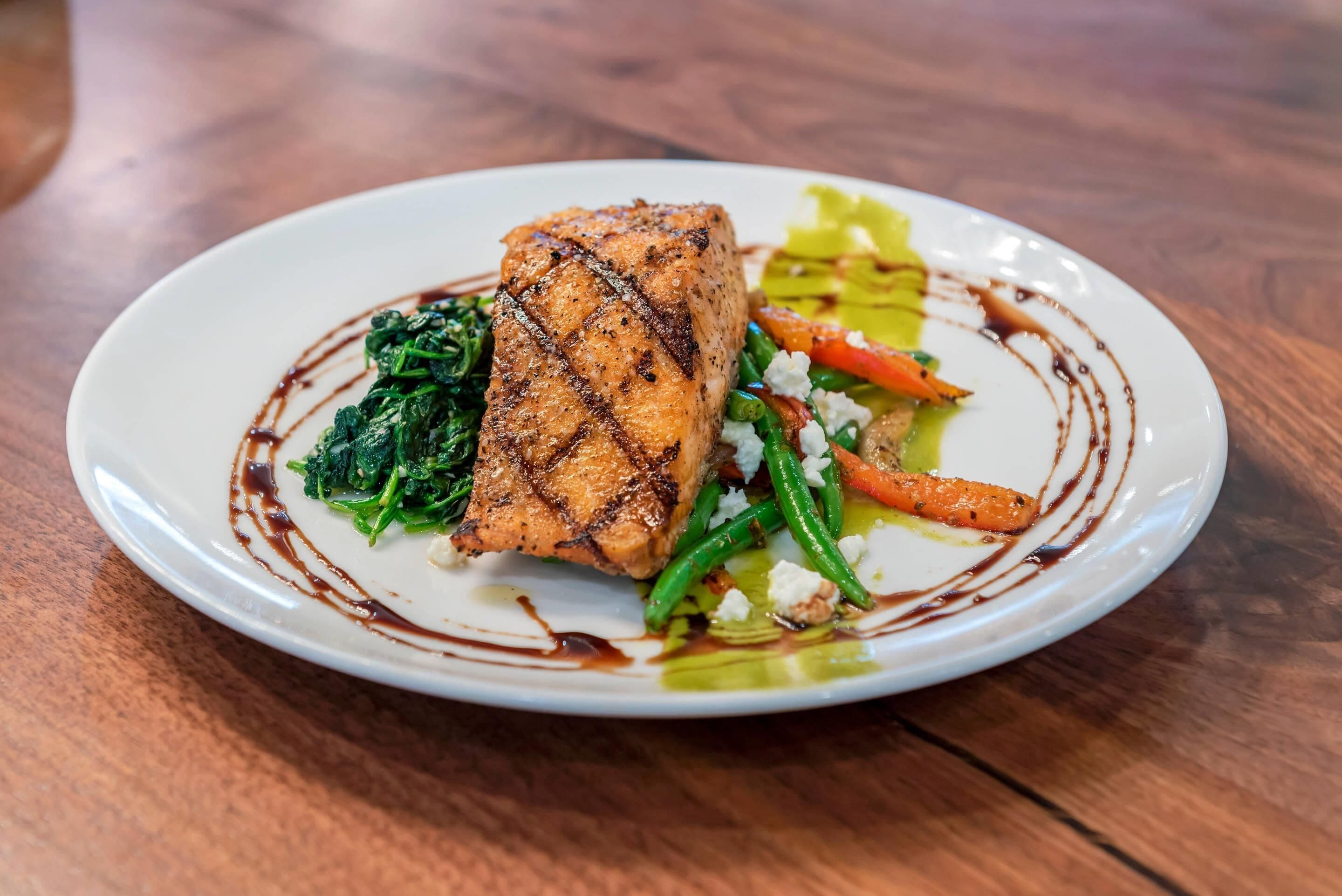 A plate of grilled salmon with vegetables and a sauce on a white plate.