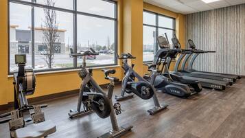 Un gimnasio con equipo de cardio, ventanas amplias y vista a un estacionamiento.