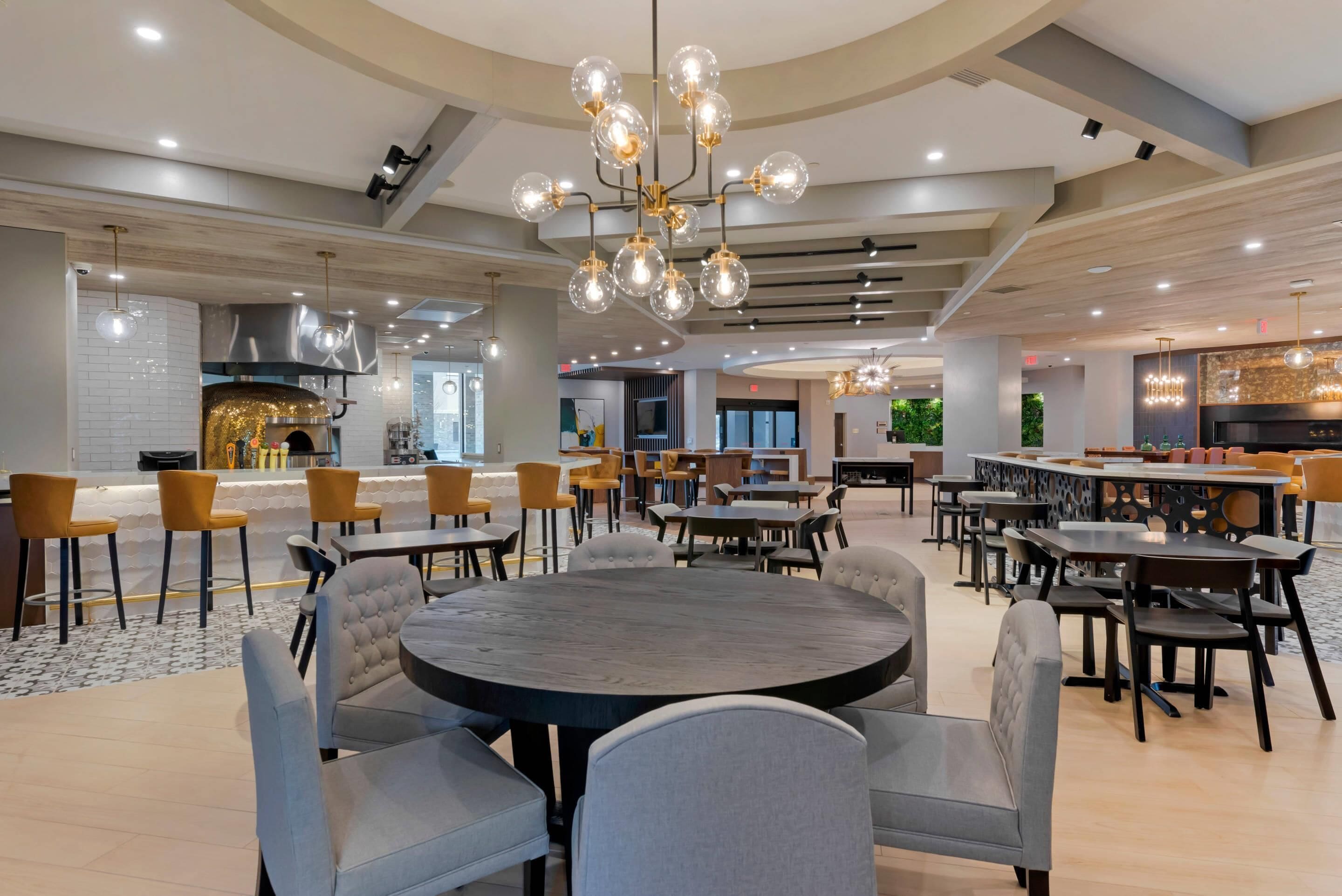 A spacious dining area with round tables and chairs, a modern chandelier, and a kitchen in the background.