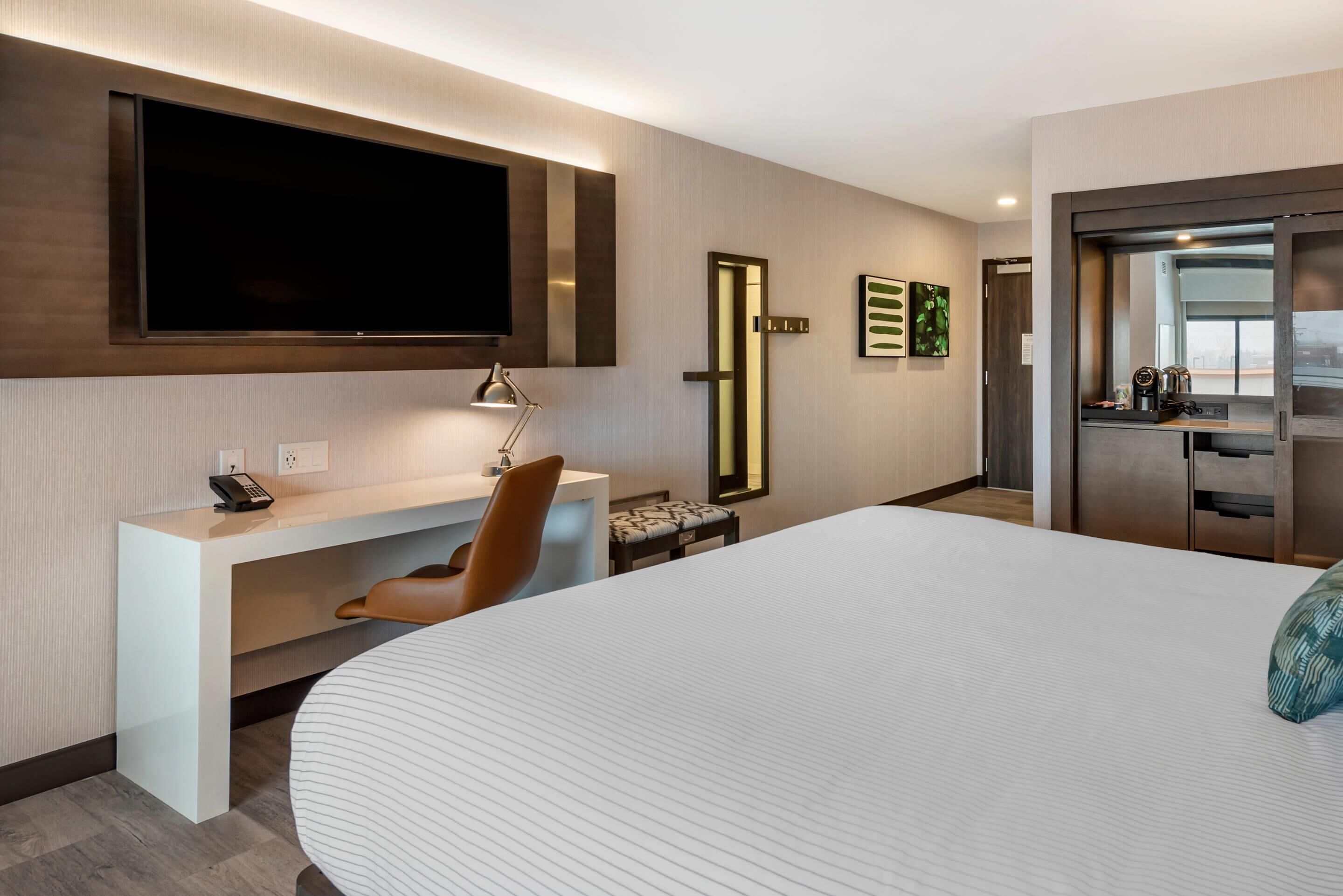Une chambre d’hôtel moderne dotée d’un grand lit, d’un bureau avec une lampe, d’un téléviseur fixé au mur et d’un petit coin cuisine.