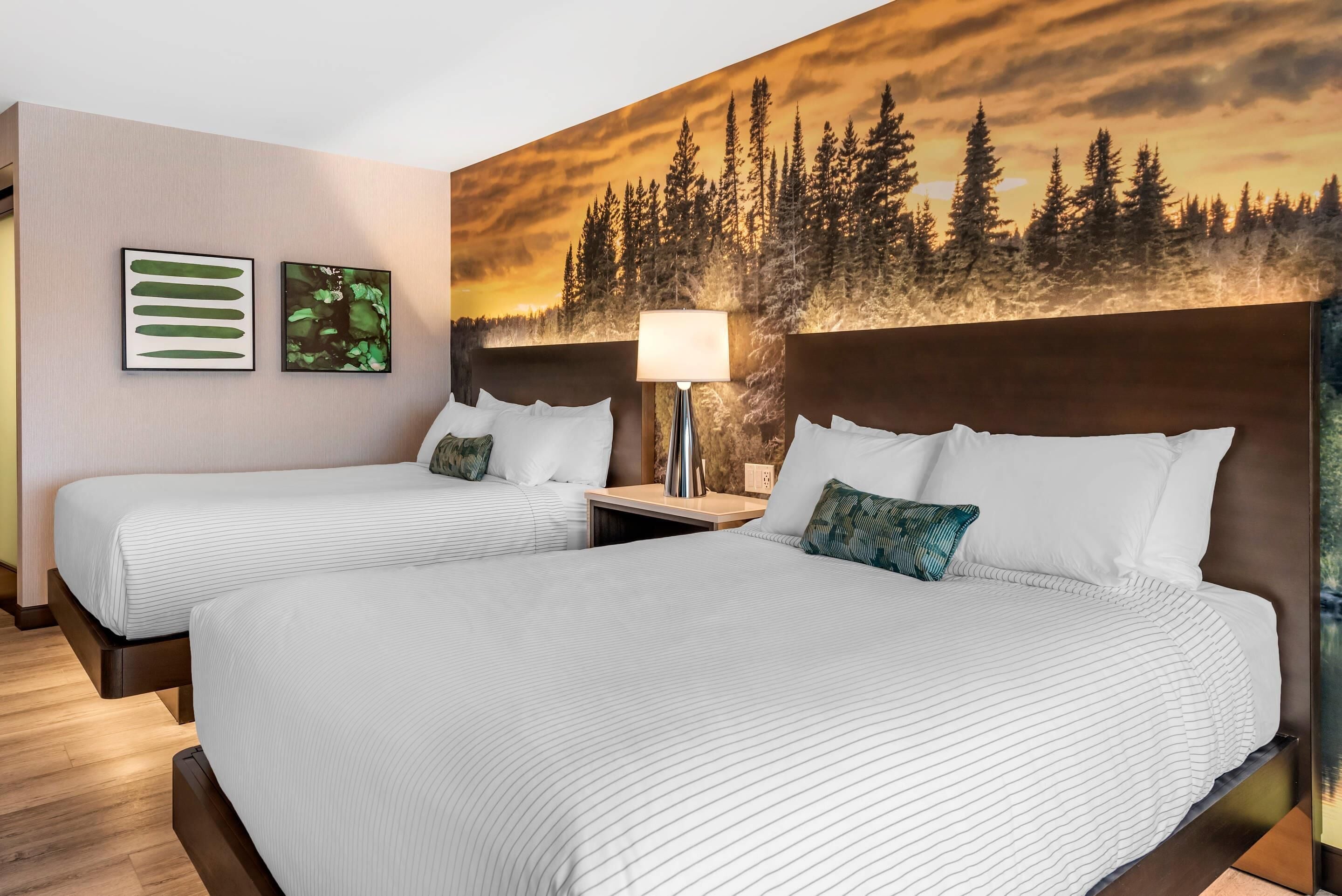 Une chambre d’hôtel avec deux lits, une table de chevet, une lampe et une grande fresque murale sur le thème de la forêt.