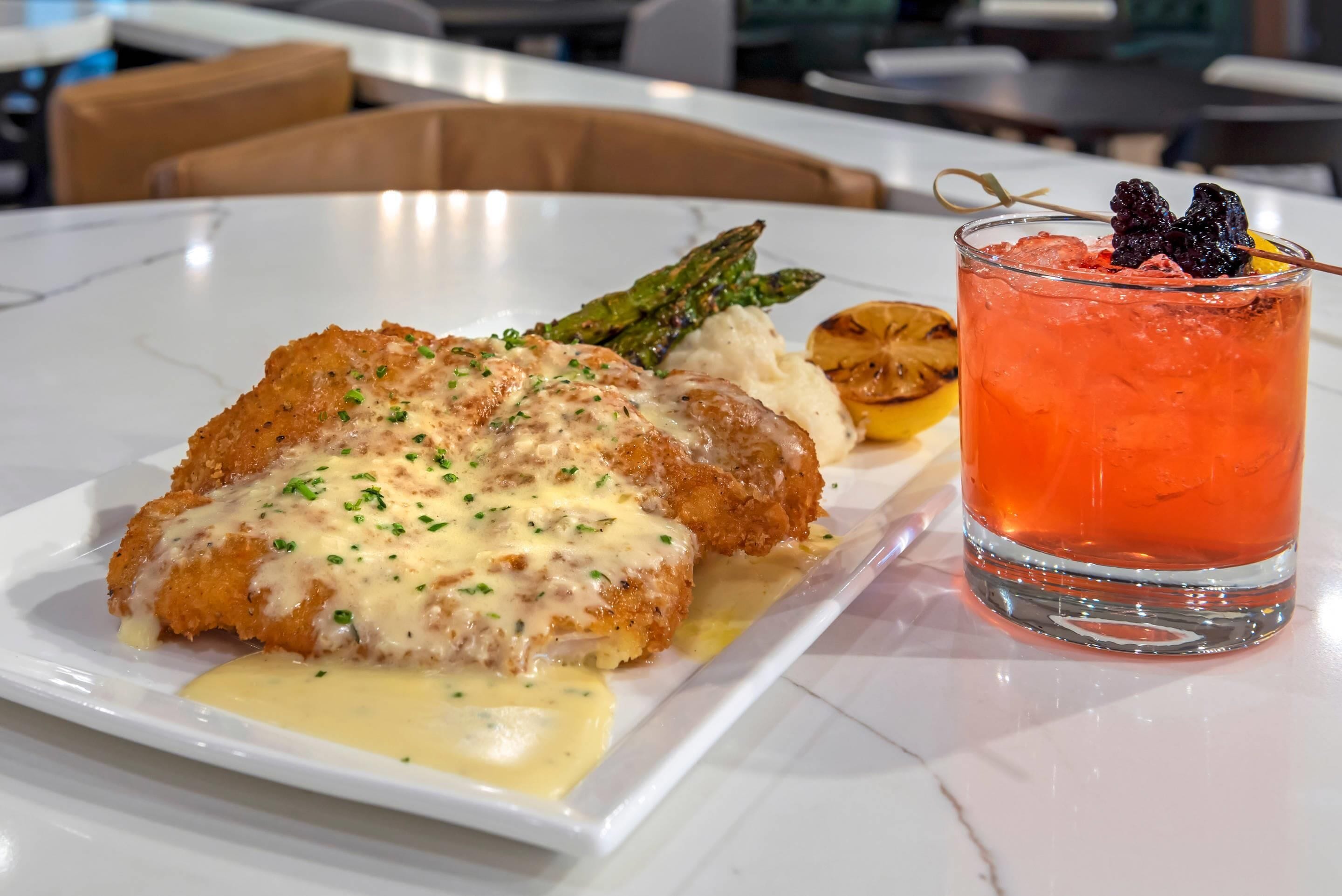 A plate of food with a breaded chicken cutlet, asparagus, and mashed potatoes, accompanied by a glass of red beverage with berries.