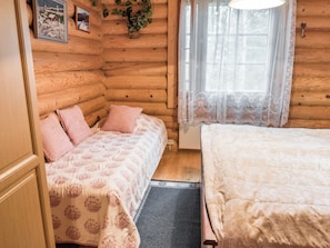 Une chambre confortable de style cabane, avec un intérieur en bois, un lit recouvert d’une courtepointe à motifs floraux, une fenêtre ornée de rideaux légers et une petite plante accrochée au mur.