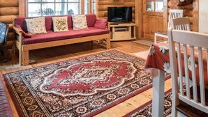 A cozy log cabin interior with a wooden sofa, a television, a dining table with chairs, and a patterned rug.
