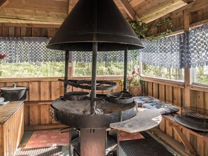 A wood-paneled room with a central wood-burning stove, a window with lace curtains, and a view of greenery outside.