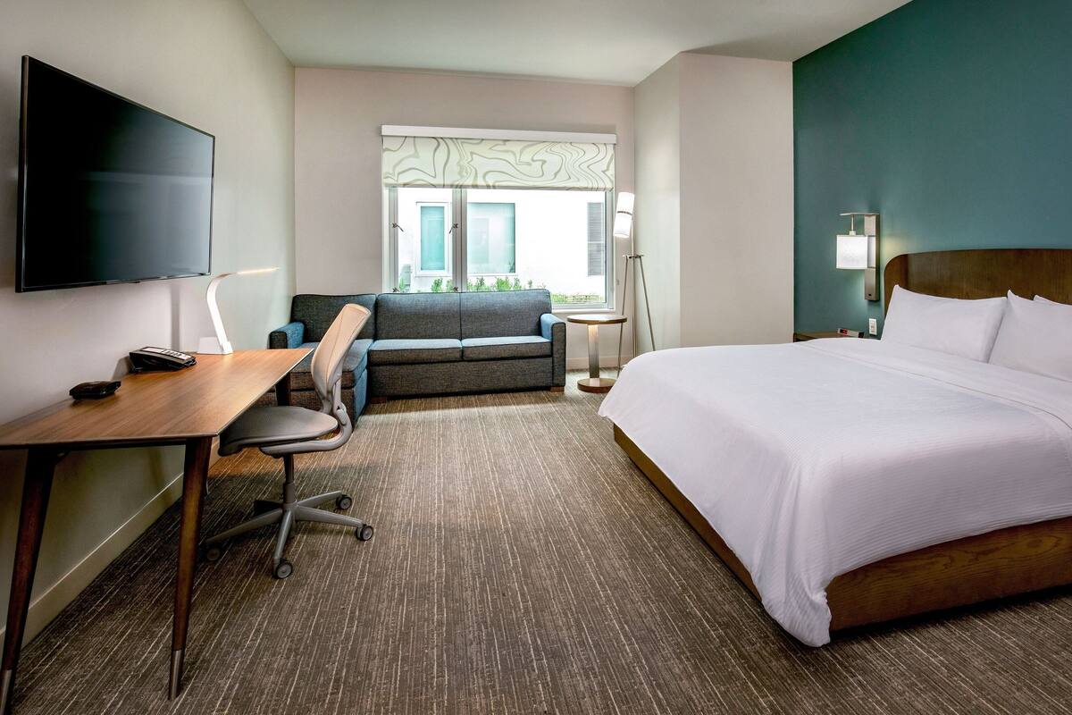 A hotel room with a bed, a desk, a chair, a TV, a window with a view, and a lamp.
