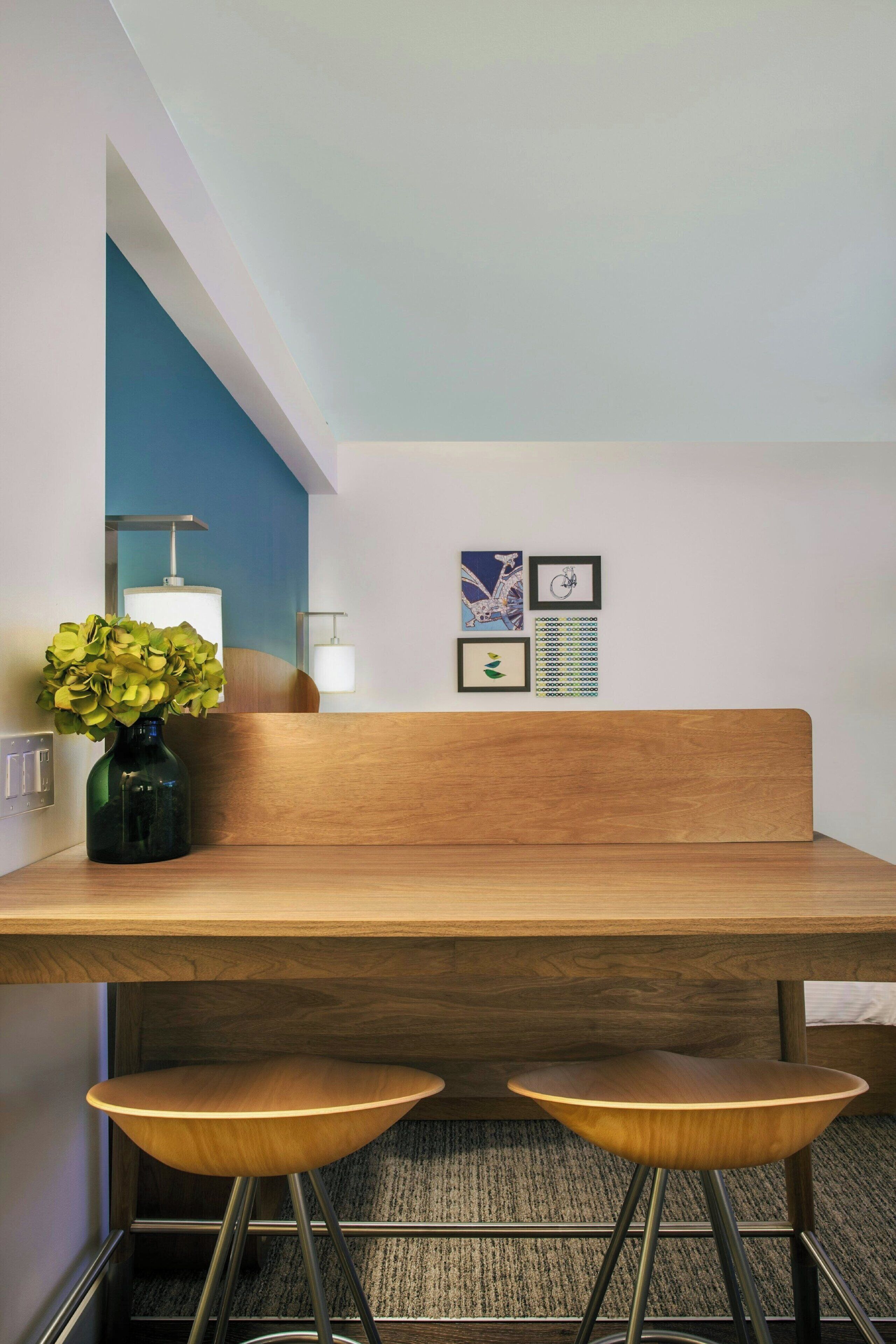 A modern room with a wooden bench and two stools, a vase with green plants, and framed artwork on the wall.