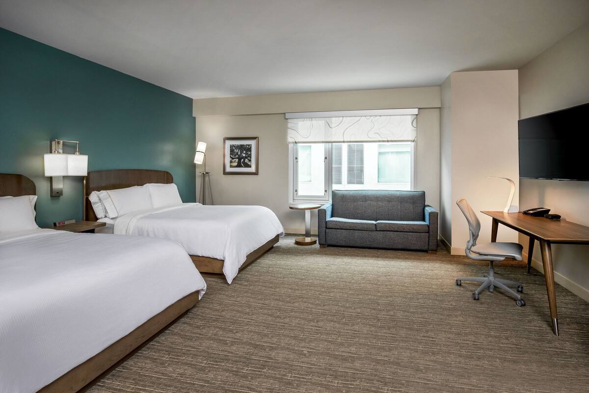 A hotel room with two beds, a desk, a chair, a TV, and a window with blinds.