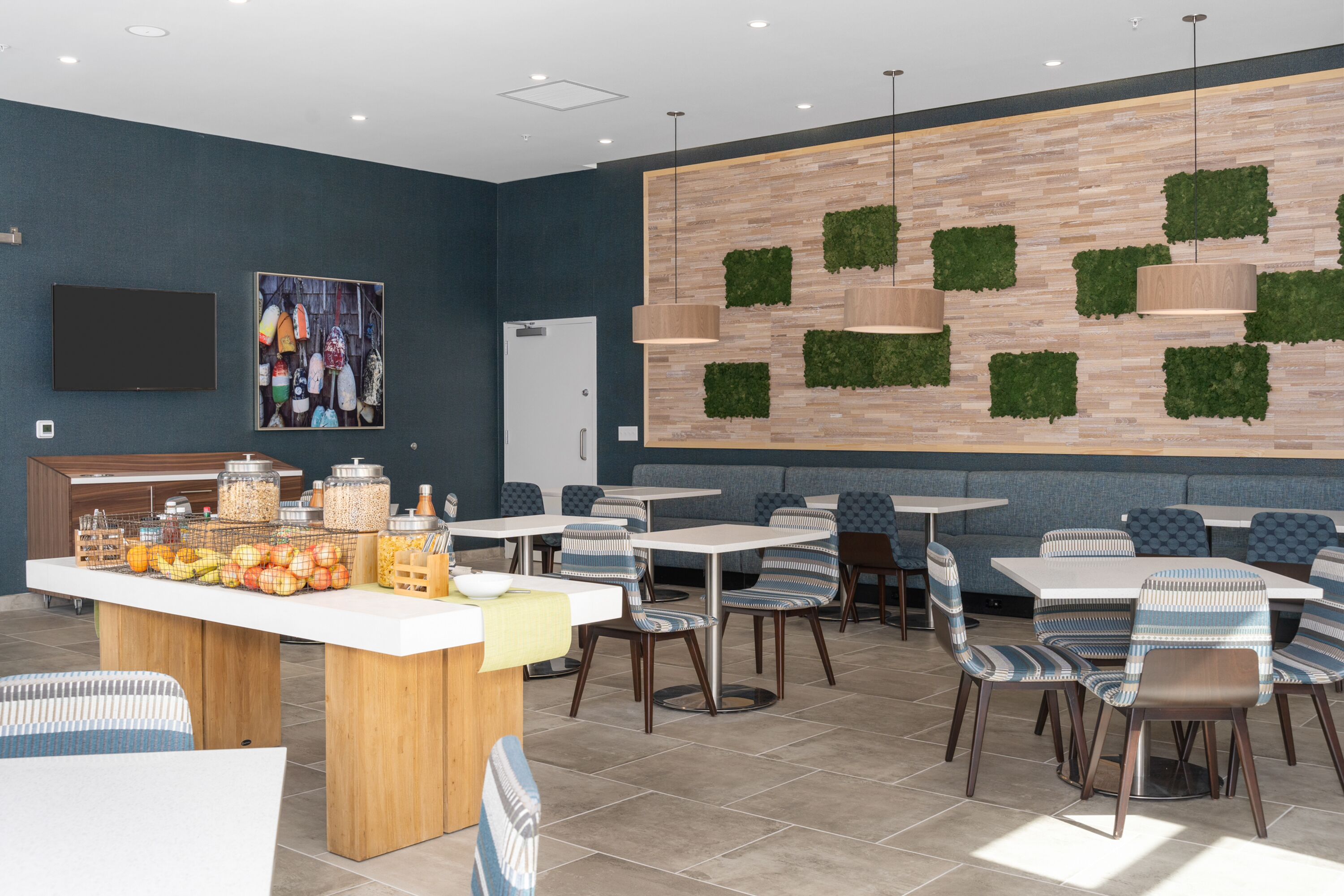 A modern restaurant with a counter displaying fruits and condiments, seating with striped chairs, and a wall with green planters.