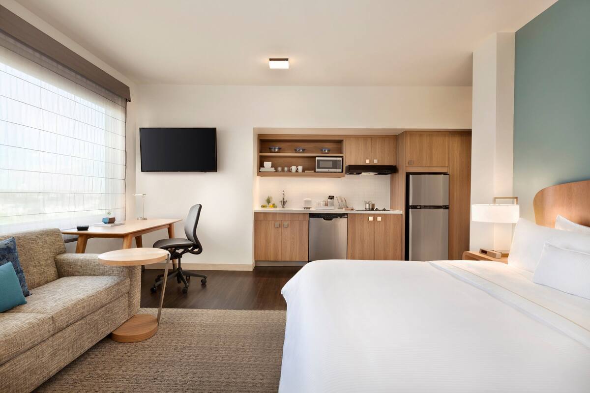 A hotel room with a bed, a sofa, a desk, a chair, a TV, a kitchen area with a fridge, microwave, and cabinets.