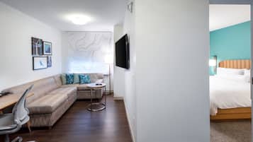 Ein modernes Hotelzimmer mit Ecksofa, einem kleinen runden Tisch, einem an der Wand befestigten Fernseher und einem Bett mit Kopfteil.